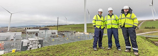Three engineers from WPD stand pictured with cloudy skies and windmills in background.