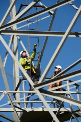 Two fieldworkers in yellow jackets climb a transmission tower