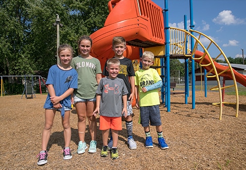 Group of children varying in age from 4-8 pose in front of an orange playground on sunny day