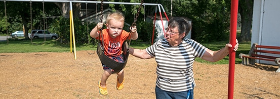 Grandmother in gray striped shirt pushing her blonde toddler grandson on a swing