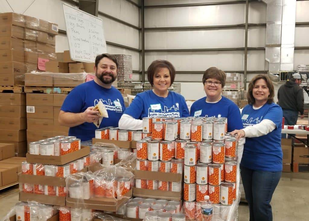 Four ppl employees in blue shirts pose with large stacks of cans at food bank
