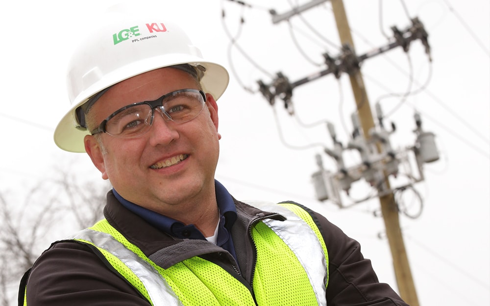 A man in an LG&E and KU hard hat and safety vest smiles in front of electric distribution poles