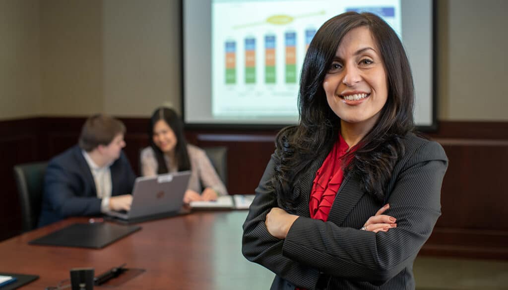 A Hispanic woman in a black suit and red top stands in front of a financial presentation.