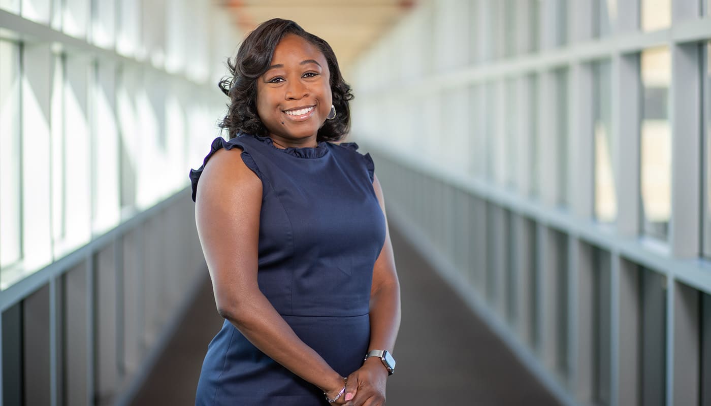 A Black woman smiles at the camera in a hallway of windows.