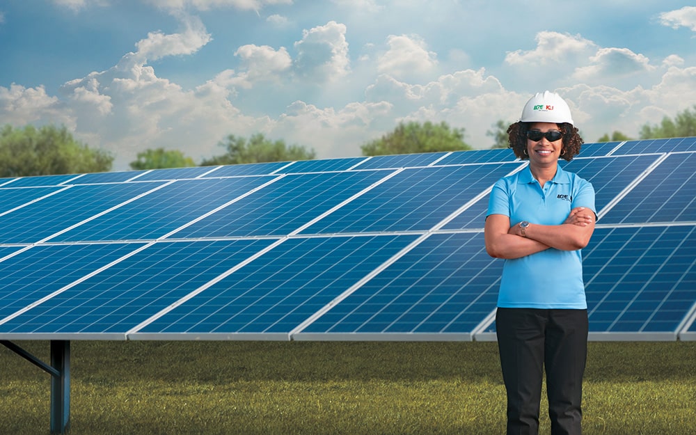 Woman in a blue shirt and hard hat standing in front of a solar panel and sunny blue sky.