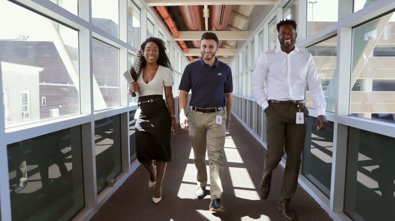A female and two male office workers walking through a glass hallway.