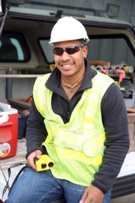 A smiling Black male wearing a hard hat and yellow safety vest sits on the back of a work truck.